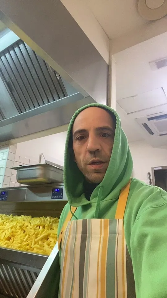 Raffaele con la felpa verde e il grembiule a righe in cucina, accanto a una vasca di patatine