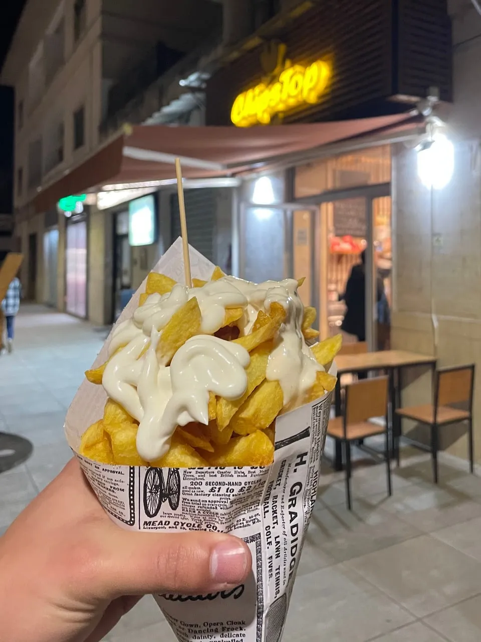 Un cono di patatine con la maionese, nella carta stampata a giornale, davanti all'insegna accesa di ChipsTop di sera